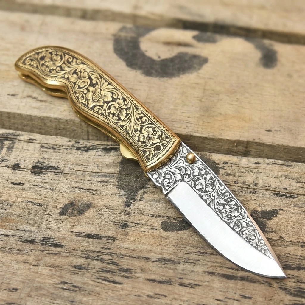 Handmade folding pocket knife with detailed floral engravings on the blade and brass handle, displayed on a weathered wooden plank surface