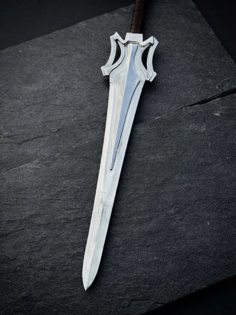 Steel blade of a handmade He-Man Power Sword featuring a distinctive flared guard, lying flat on a dark slate stone surface