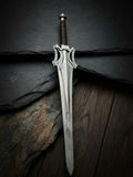 Handmade He-Man Power Sword replica featuring a steel blade, iconic flared crossguard, and brown leather-wrapped handle resting on black slate and wood surfaces
