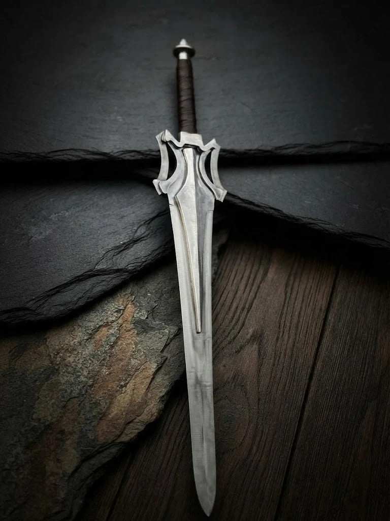 Handmade He-Man Power Sword replica featuring a steel blade, iconic flared crossguard, and brown leather-wrapped handle resting on black slate and wood surfaces