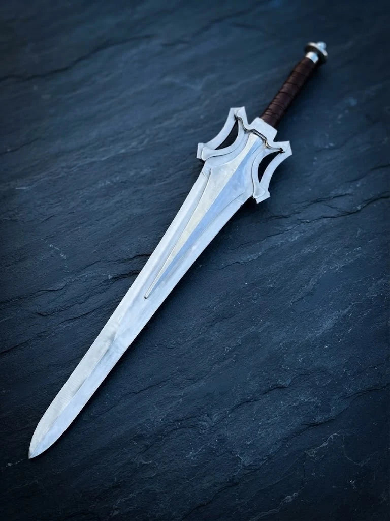 Full-length view of a handmade He-Man Power Sword replica lying diagonally on a textured dark slate stone background, featuring a steel blade and leather-wrapped grip