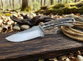 A custom handmade cowboy belt knife forged from a steel rasp file, featuring a drop-point blade with prominent cross-hatched rasp teeth marks and a textured stag antler horn handle with a green lanyard hole lining, resting on a rustic wooden log with a coiled rope and an outdoor forest background.