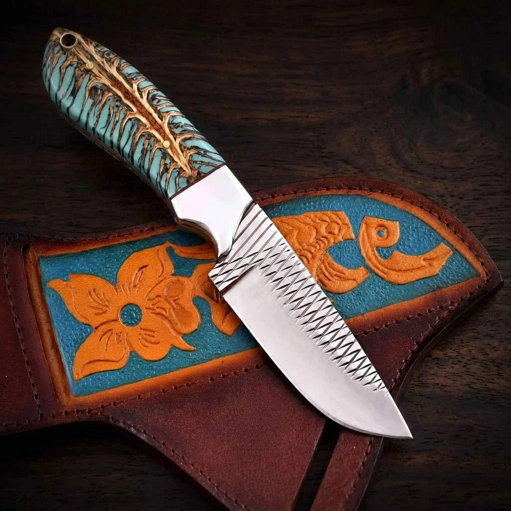 A handmade rasp steel cowboy knife featuring distinct cross-hatched file marks on its blade and a turquoise-dyed pine cone hybrid resin handle, placed diagonally over its custom tooled leather crossdraw sheath with orange floral designs against a dark wood grain backdrop.