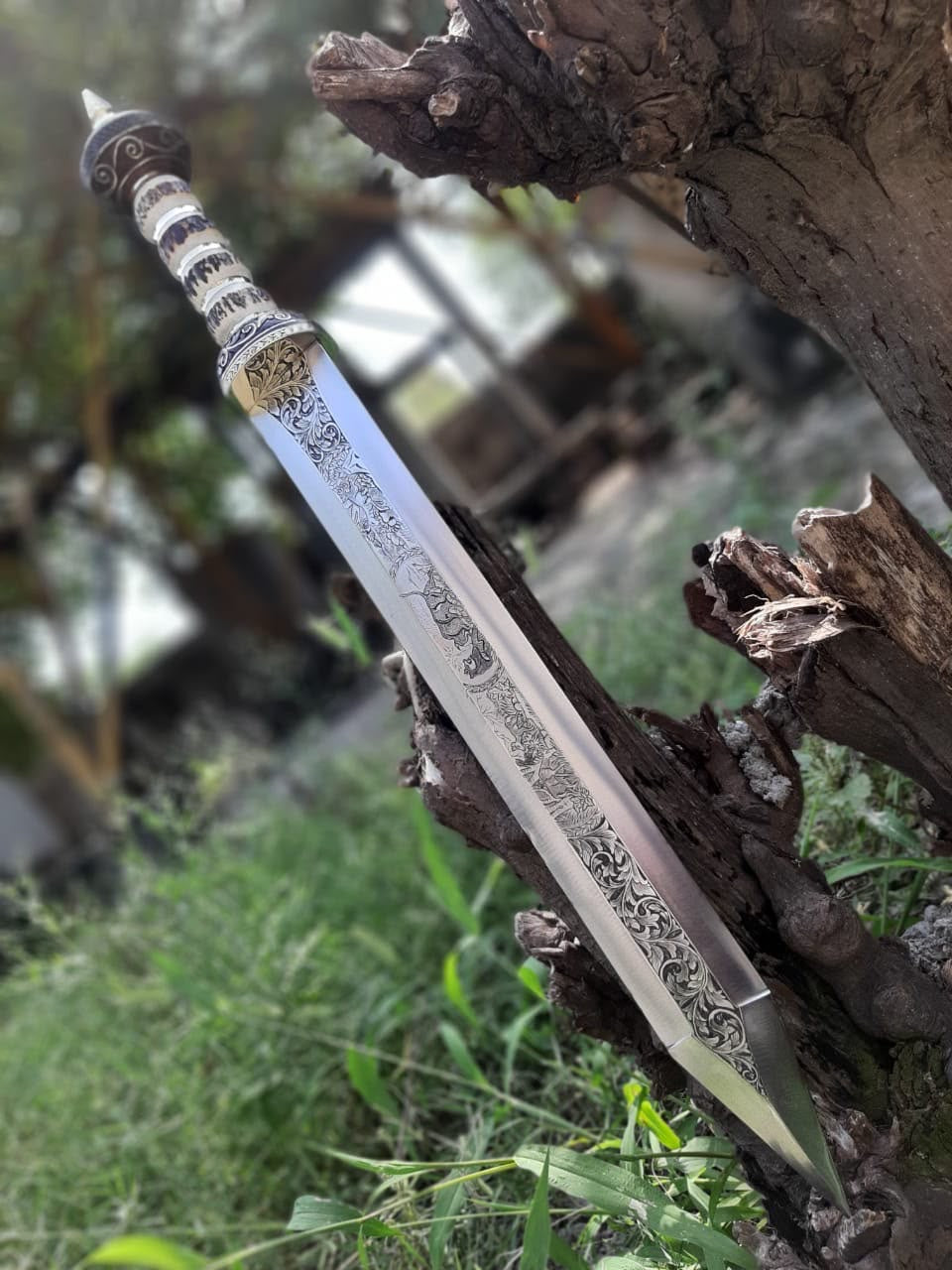 Handmade Roman Gladius short sword featuring an intricately engraved blade and carved bone handle, displayed outdoors resting against natural wood driftwood.