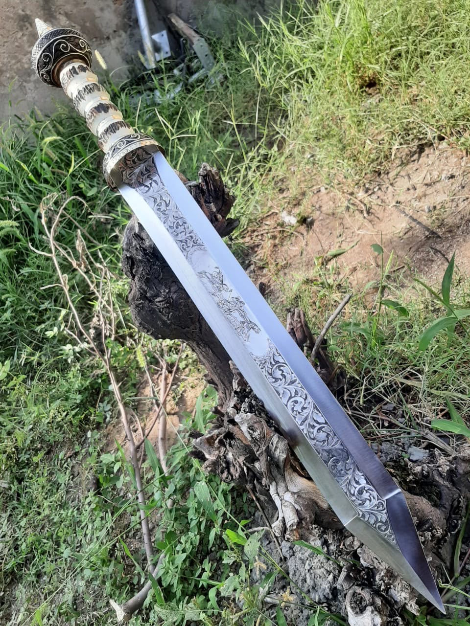 Full-length outdoor view of a handmade Roman Gladius short sword with an intricately engraved blade and carved bone handle leaning against wood