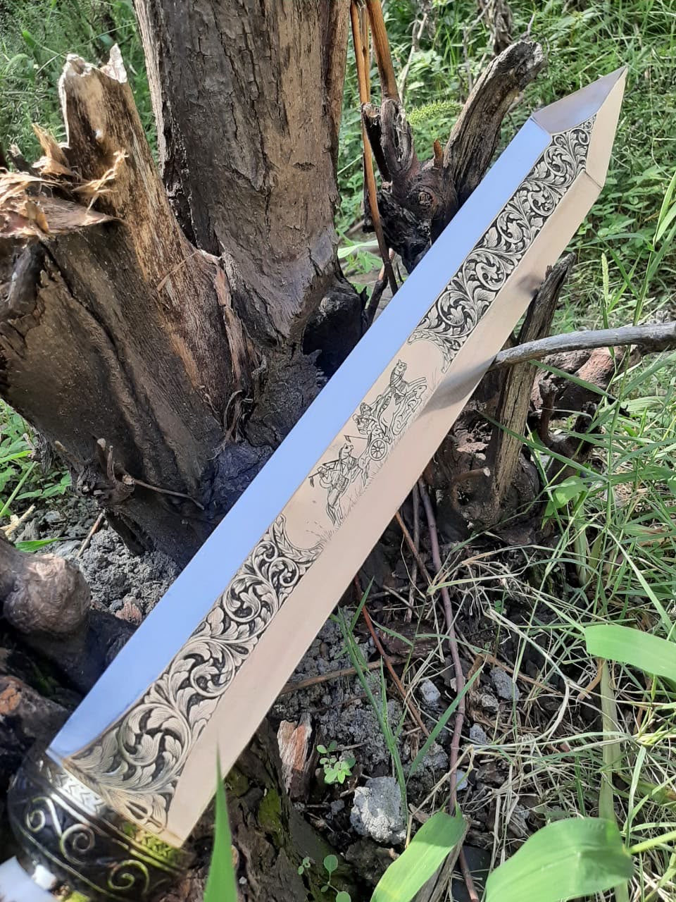 Close-up view of a Roman Gladius short sword blade featuring detailed floral engravings and a medieval chariot scene motif, outdoors against natural wood