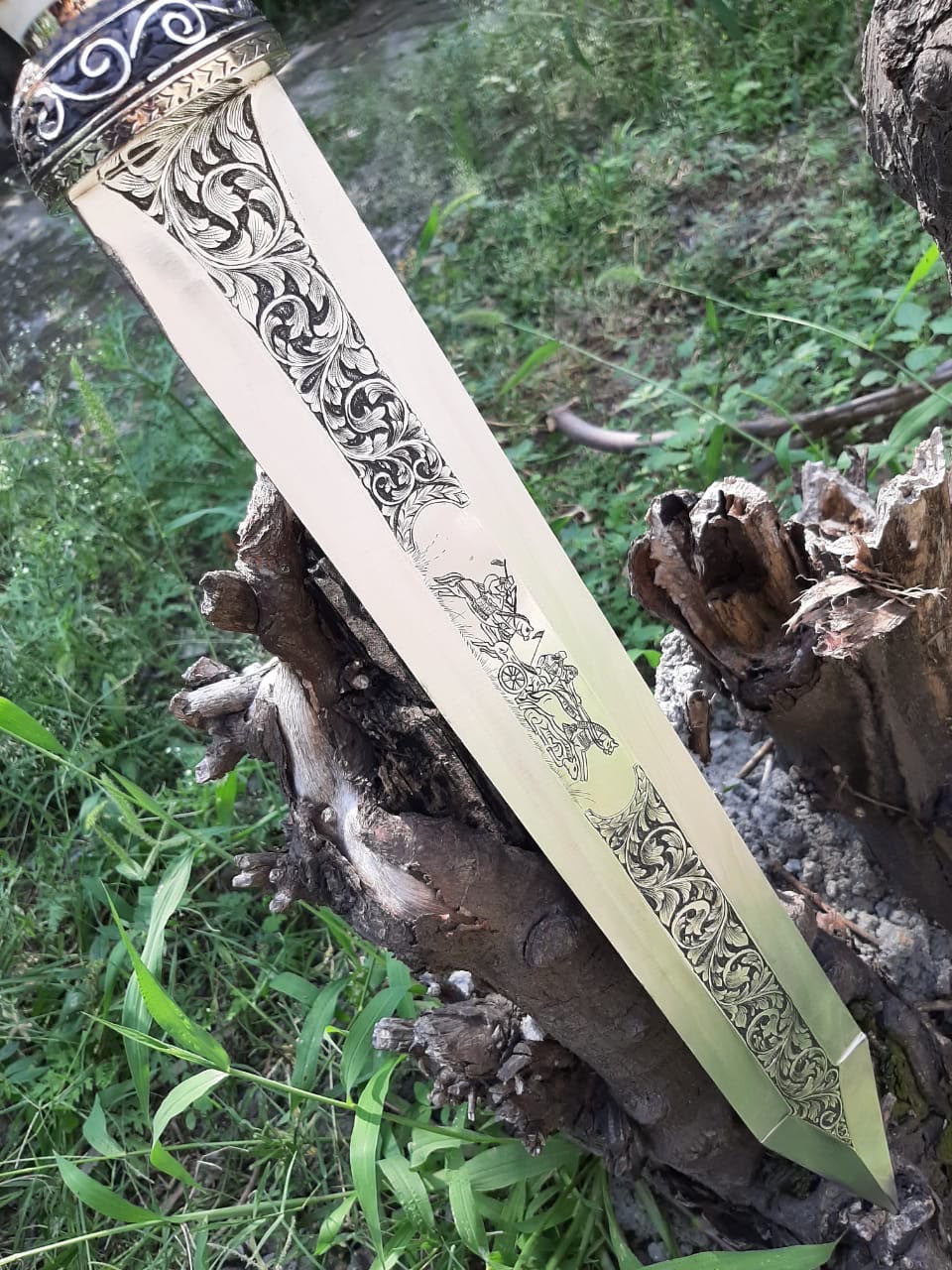 Detailed view of the reverse side of a Roman Gladius short sword blade, highlighting ornate floral scrollwork and a chariot scene engraving against an outdoor natural wood backdrop.