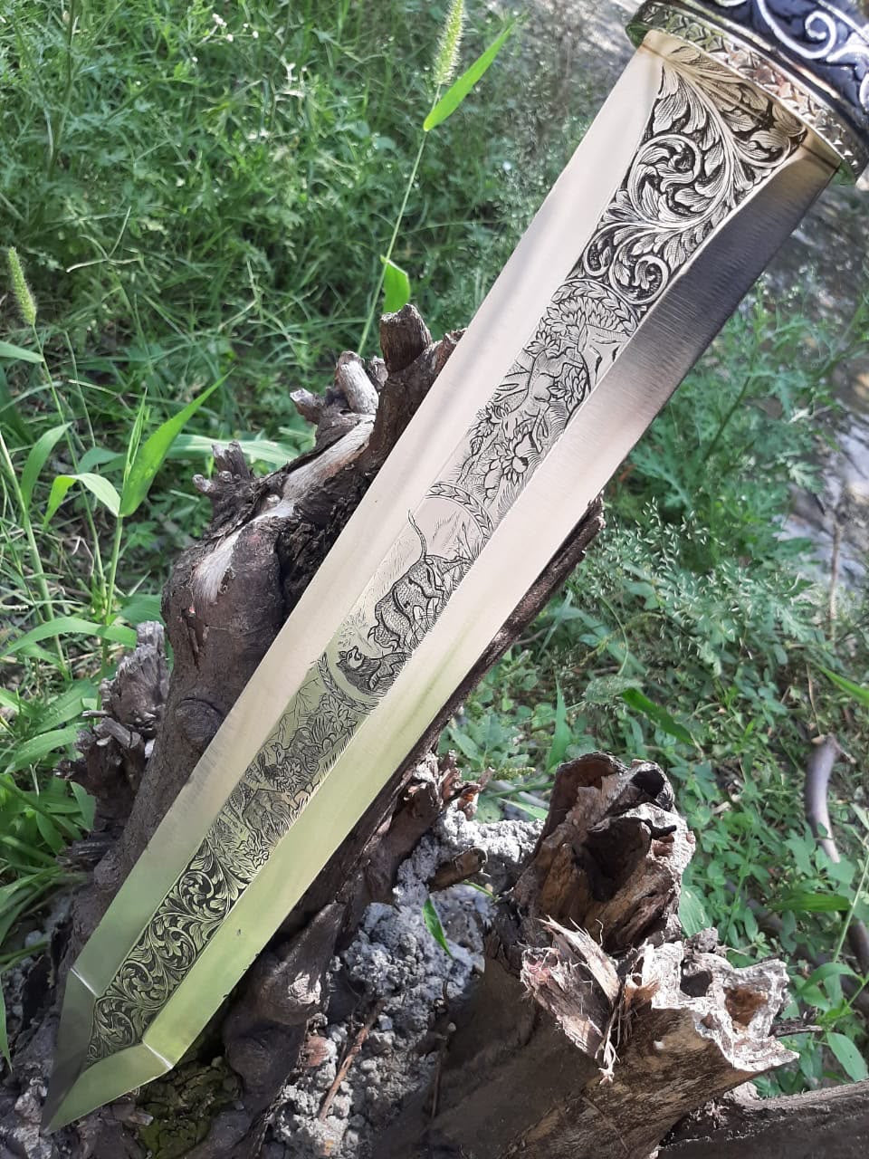 Close-up view of a Roman Gladius short sword blade featuring intricate wildlife and floral engravings, shown outdoors leaning against natural wood driftwood