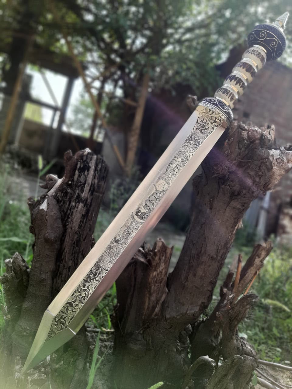Full upright view of a handmade Roman Gladius short sword featuring an ornate engraved blade and stag bone handle resting against a tree stump outdoors.