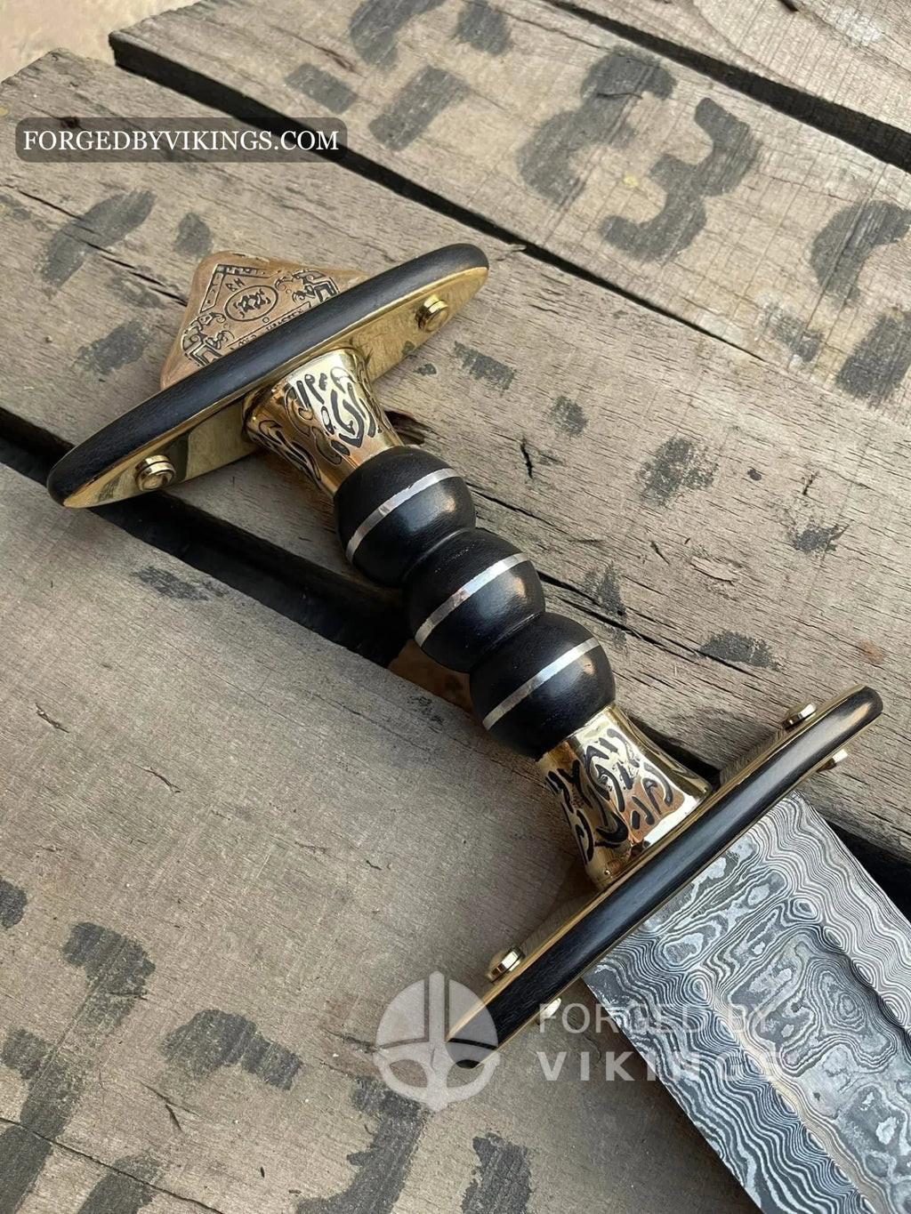 Detailed close-up shot of a Viking sword handle featuring a lobed pommel, engraved brass accents, black ribbed wooden grip, and crossguard resting on wooden crates.