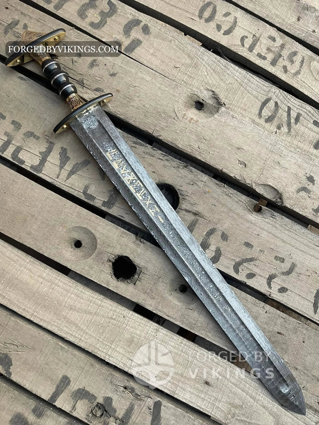 Handmade Viking sword with a pattern-welded Damascus steel blade inscribed with runes, featuring a ornate brass and dark wood hilt resting diagonally on a wooden crate surface