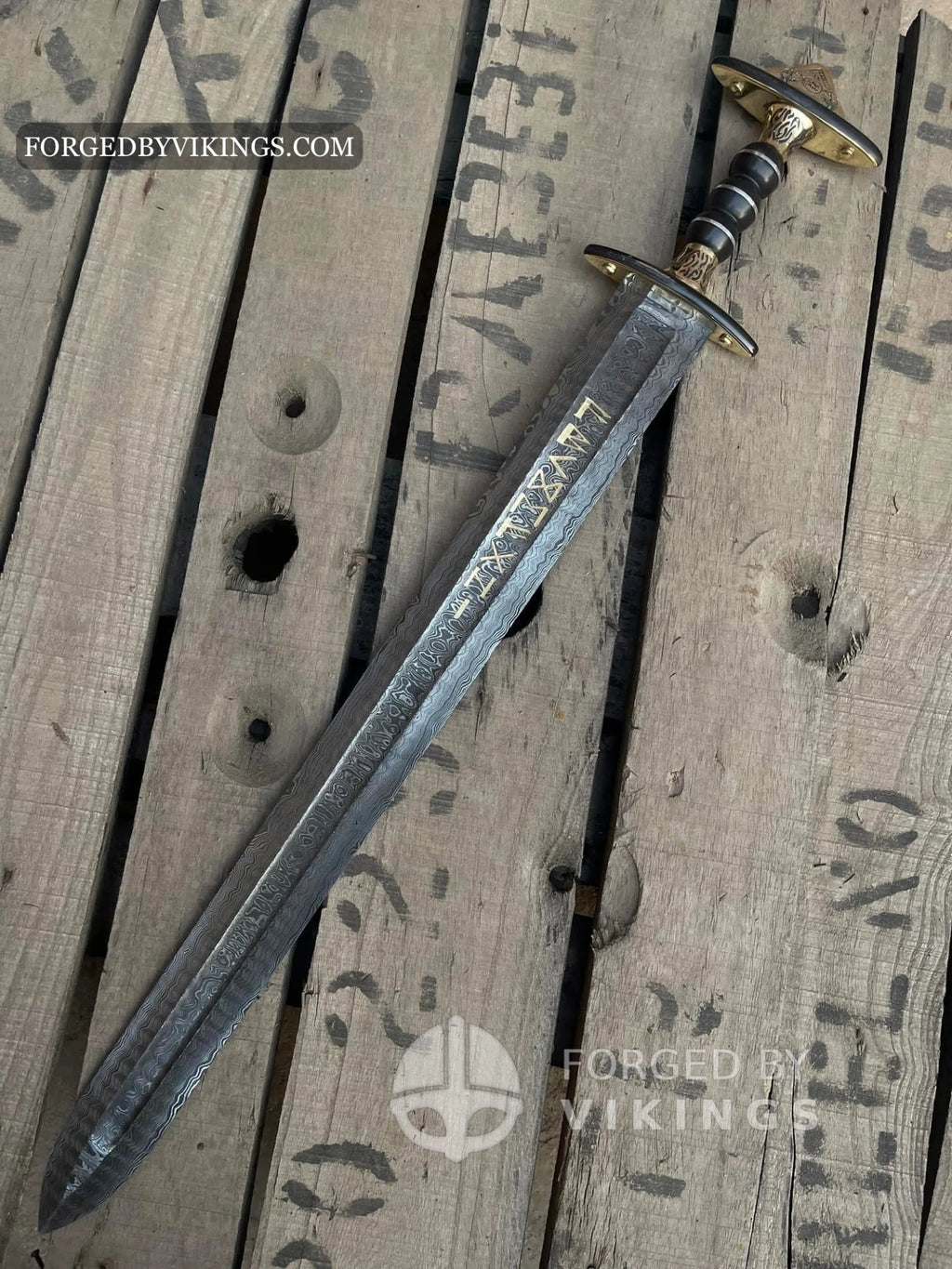 Handmade Viking sword featuring a Damascus steel blade with gold runic inscriptions, detailed brass crossguard, and dark ribbed handle lying on a wooden crate surface