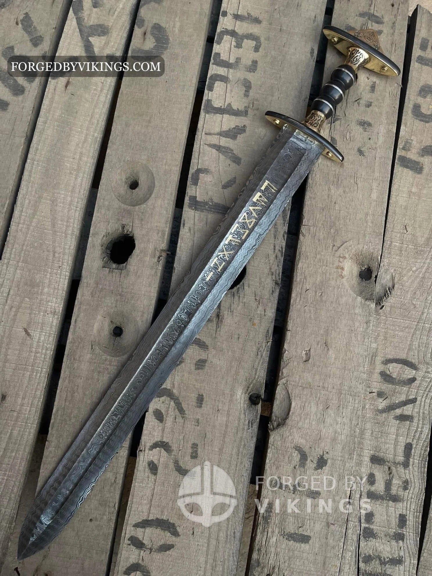 Handmade Viking sword featuring a Damascus steel blade with gold runic inscriptions, detailed brass crossguard, and dark ribbed handle lying on a wooden crate surface