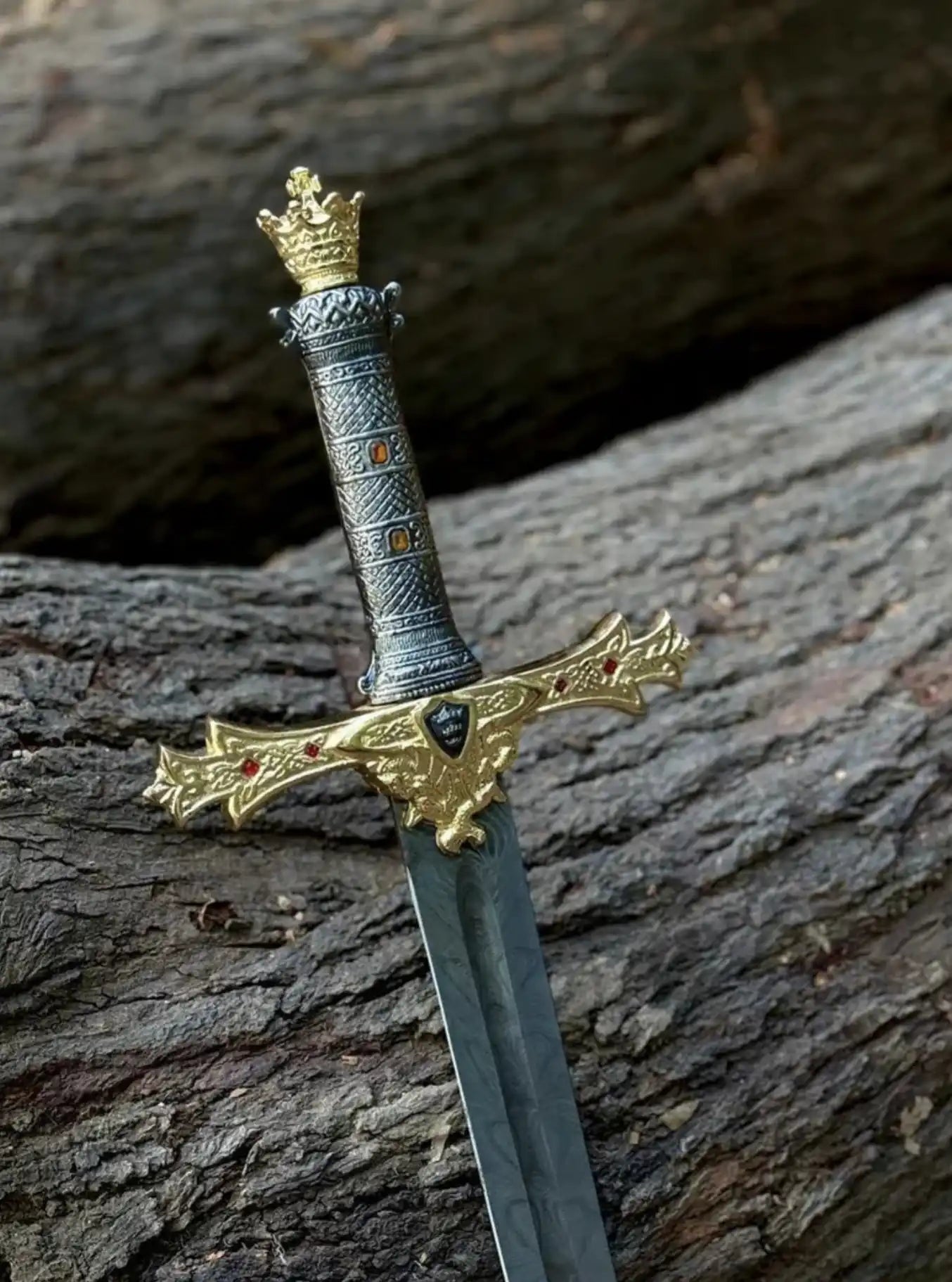 Close-up of a royal medieval sword hilt showing a golden crown pommel, a textured silver and gold handle with inset amber gems, and an ornate gold crossguard with a central black shield emblem.