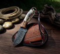 A handcrafted Damascus steel Old West cowboy knife with a patterned blade, featuring a hybrid handle made of polished rosewood bolster and textured stag antler scale with a leather lanyard, resting on top of a basket-weave tooled brown leather pancake sheath over a dark wood table with a coiled rope in the background.