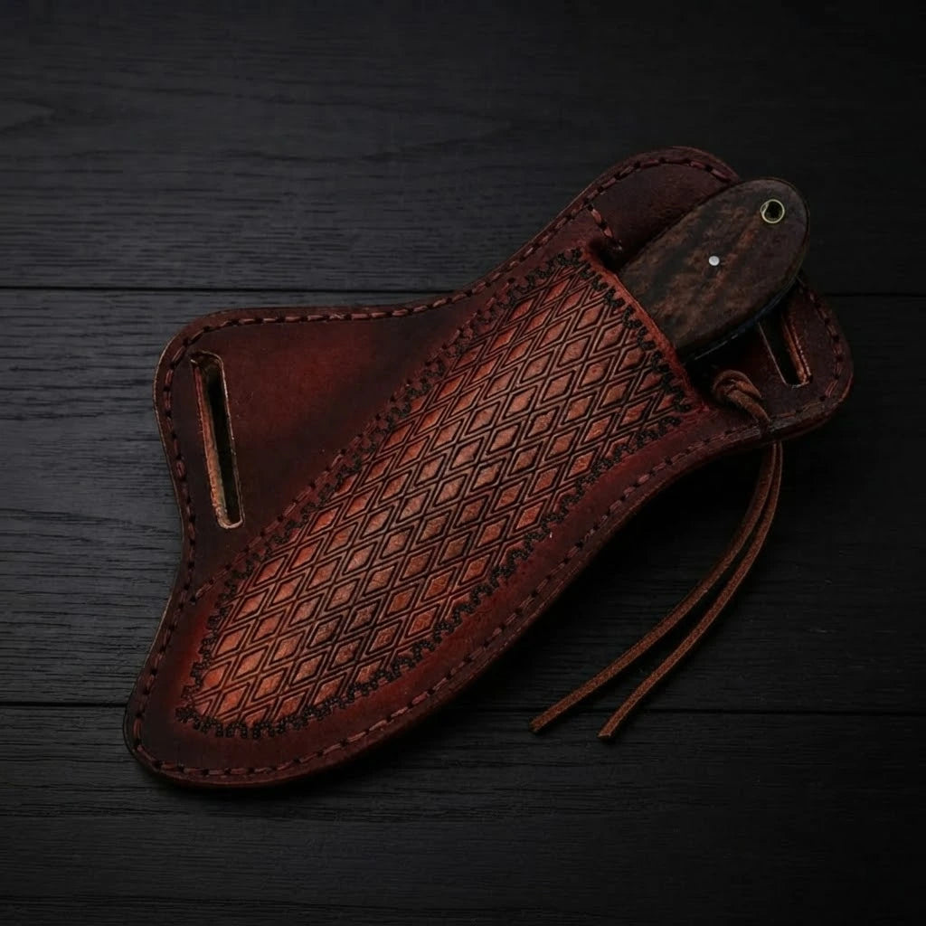 Handmade Buckaroo knife fully sheathed in a hand-tooled brown leather pancake sheath showing only the dark wood handle butt, displayed on a black wood surface