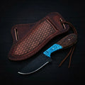 Handmade Buckaroo ranch knife crafted from dark forged rasp steel with a hybrid turquoise inlay and wood handle, shown with a textured brown leather pancake sheath on a dark wood background