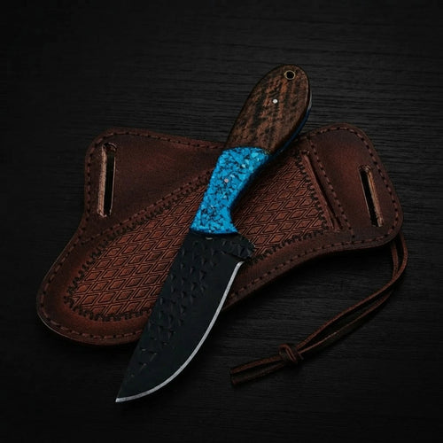 Top-down view of a handmade Buckaroo ranch knife with a dark forged rasp steel blade, turquoise inlay and wood handle, resting on a brown leather pancake sheath over dark wood.