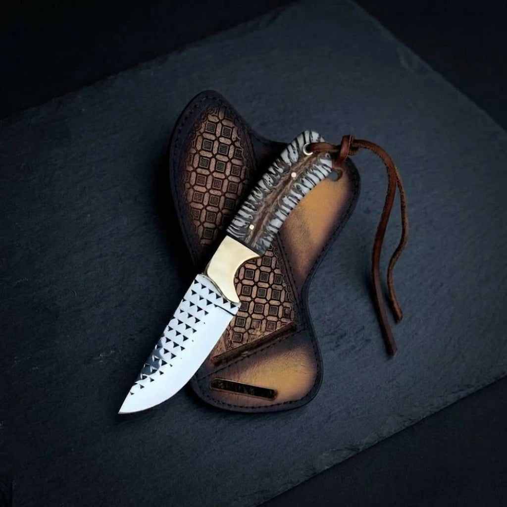 Handmade Rasp Steel Cowboy Knife with a unique cast pine cone hybrid handle, leather lanyard, and brass bolster, displayed on top of its geometrically stamped leather crossdraw sheath over a dark slate background.