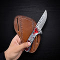 A hand holding a handmade Rasp Steel Cowboy Knife with a red and swirl resin handle in front of its stamped tan leather sheath against a dark wooden background