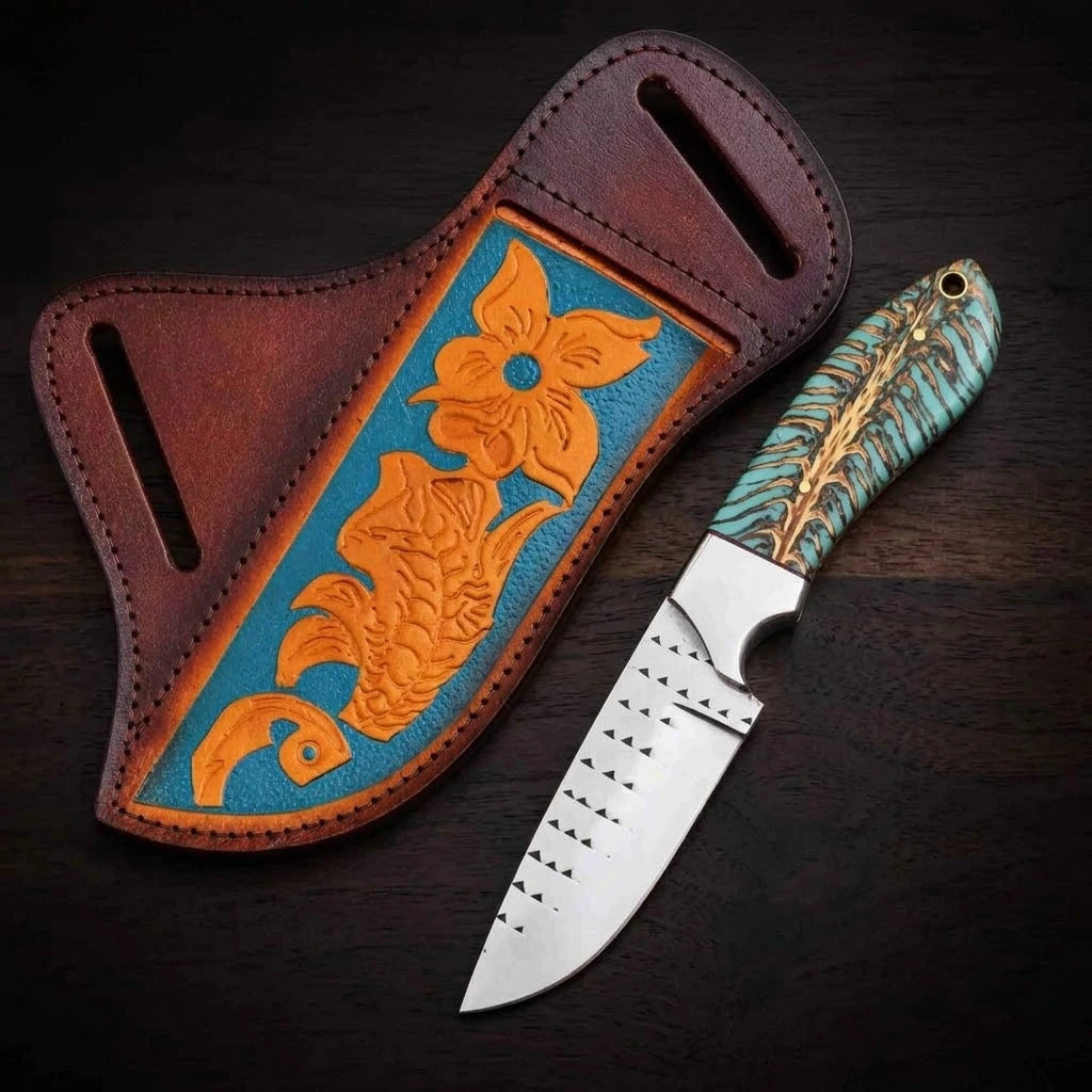 A handmade rasp steel cowboy knife featuring a turquoise-dyed pine cone hybrid handle and a steel bolster, resting next to a custom leather crossdraw sheath adorned with carved orange floral patterns on a vibrant blue background, displayed over a dark wood surface.