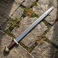 Hand-forged Viking sword with pattern-welded Damascus steel blade and brown leather hilt, laid on stone and moss. Premium authentic Norse weapon replica available for collectors in the USA, UK, and Europe.