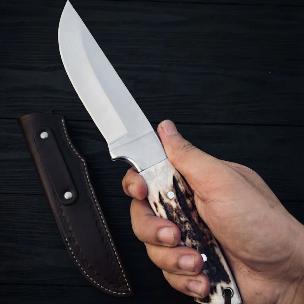 A hand holding the Stag Ridge Hunter D2 steel hunting knife, highlighting the authentic stag horn handle and steel bolster in front of a dark brown leather sheath on a black wooden surface