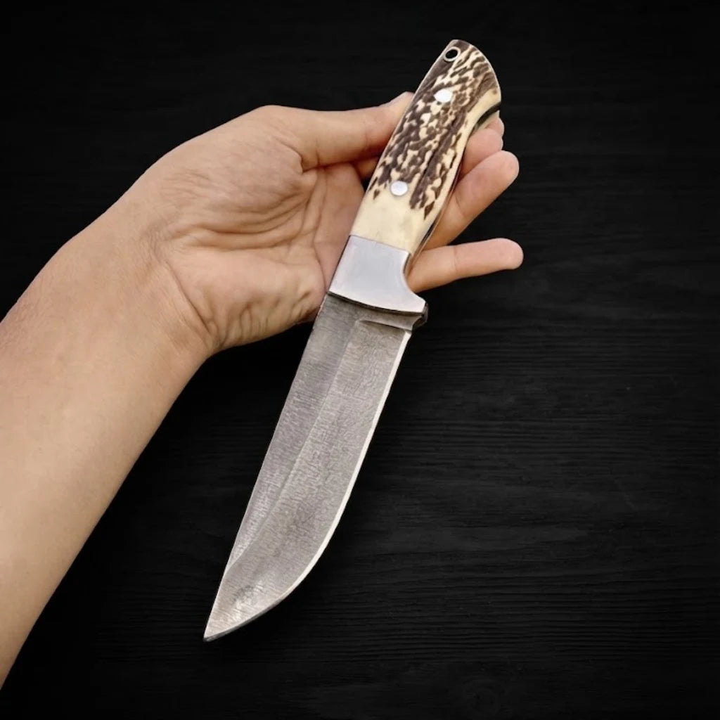 A hand holding the Stag Ridge Hunter handmade hunting knife with an authentic stag horn handle and steel blade over a dark textured wood background.