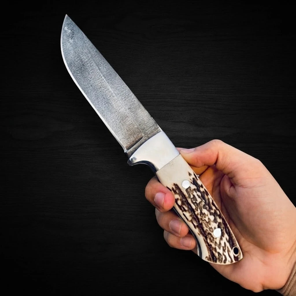 Handholding the Stag Ridge Hunter handmade hunting knife with an authentic stag horn handle, steel bolster, and textured drop point blade in front of a leather sheath