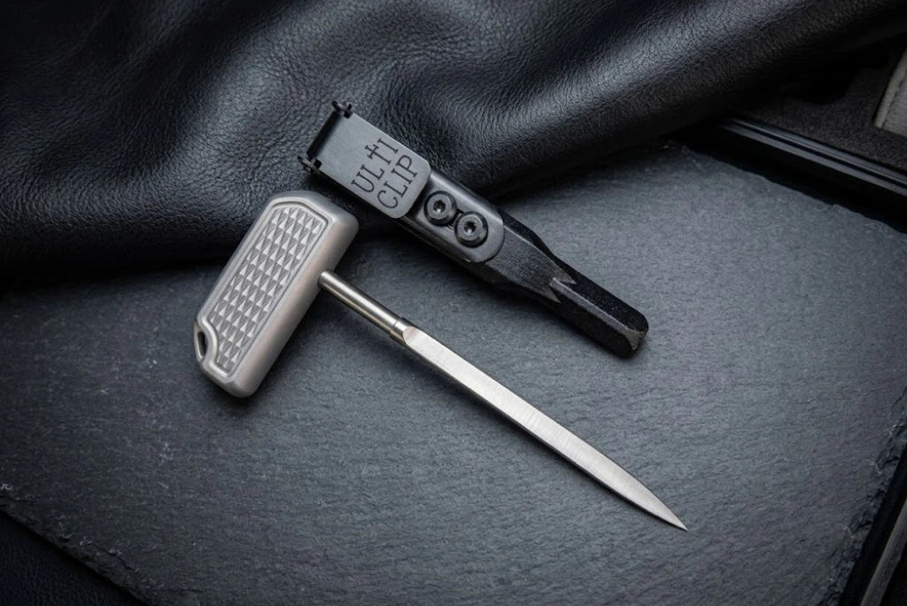 CNC machined titanium EDC push tool with a diamond-textured T-handle grip, displayed alongside its black sheath fitted with an UltiClip on a dark slate background