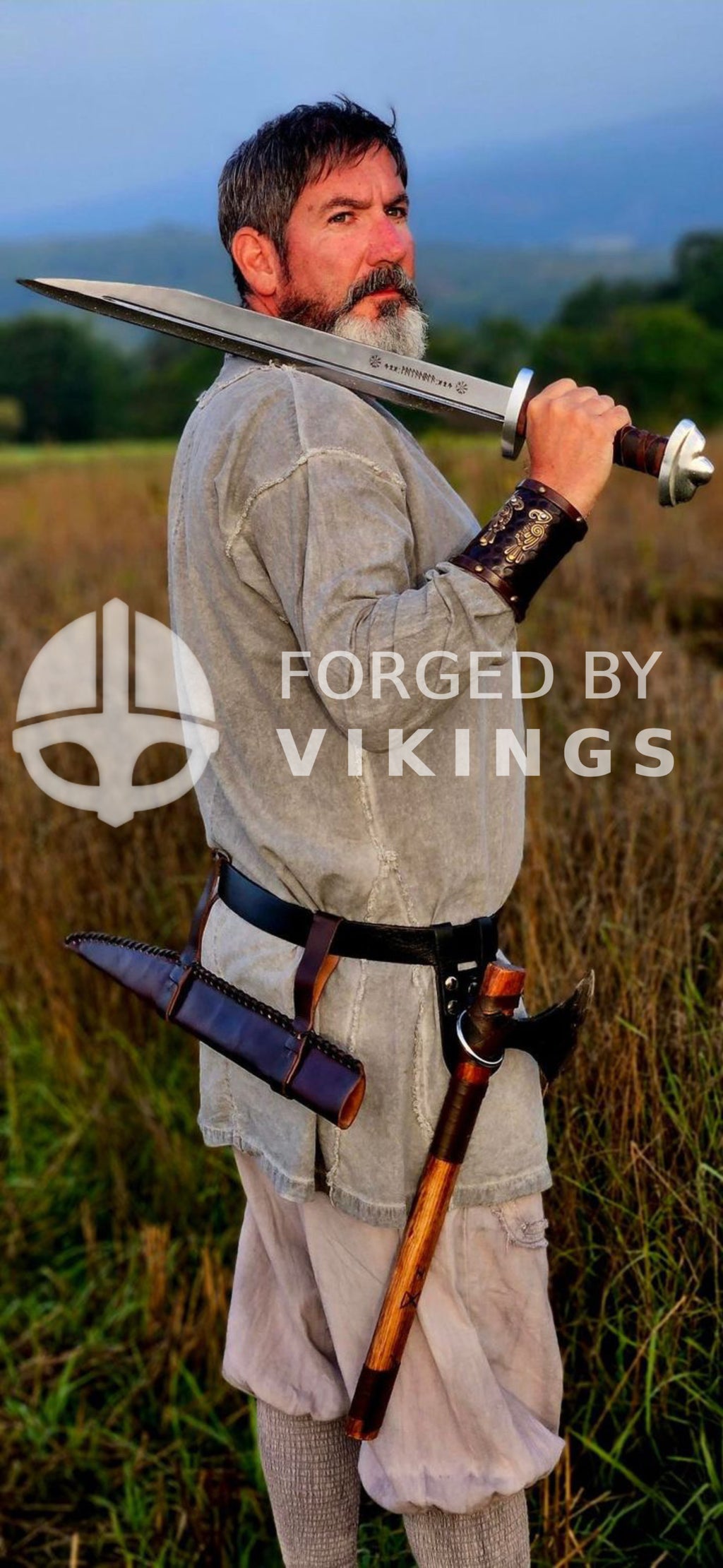 A Viking warrior dressed in historical clothing, holding a hand-forged Seax and carrying a battle axe, standing in an outdoor field.